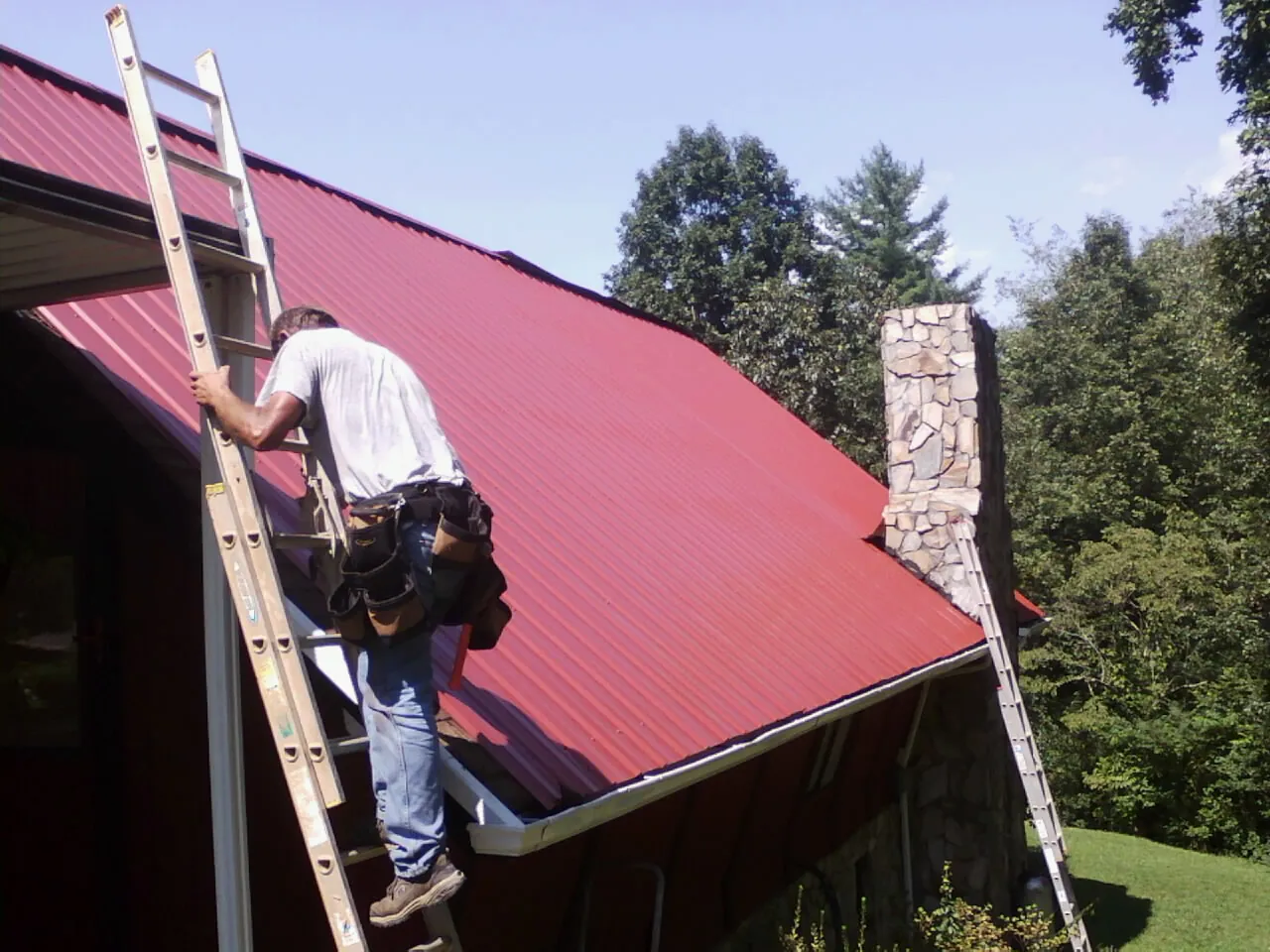 Roofer on a ladder beside a red metal roof and stone chimney — Soffit & Fascia Repair by Roofing Brothers in Miles City